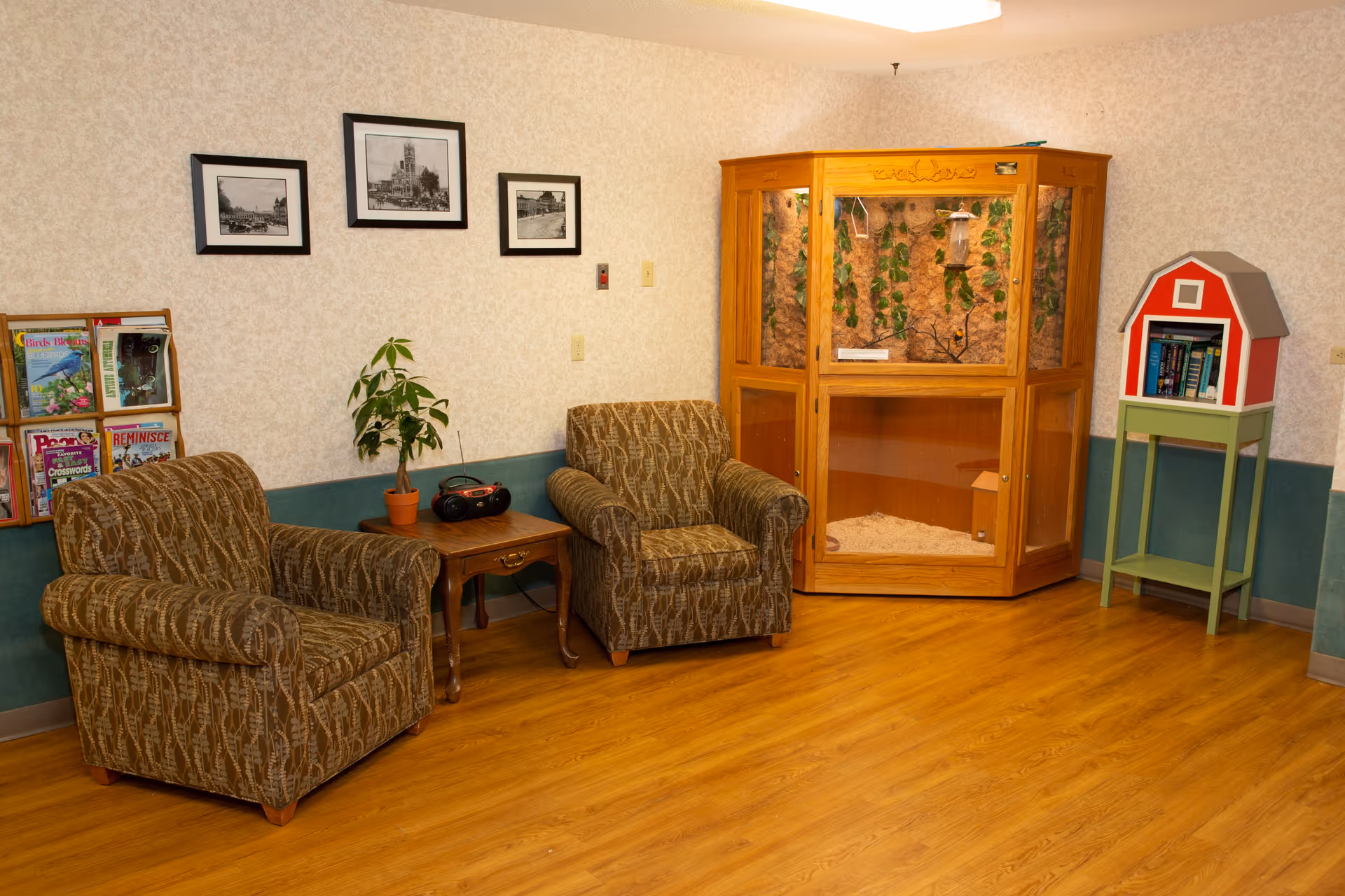 A cozy seating area with two patterned armchairs, a side table with a plant and radio, an enclosed wooden habitat/display, and a small red-barn bookshelf against a wallpapered wall.