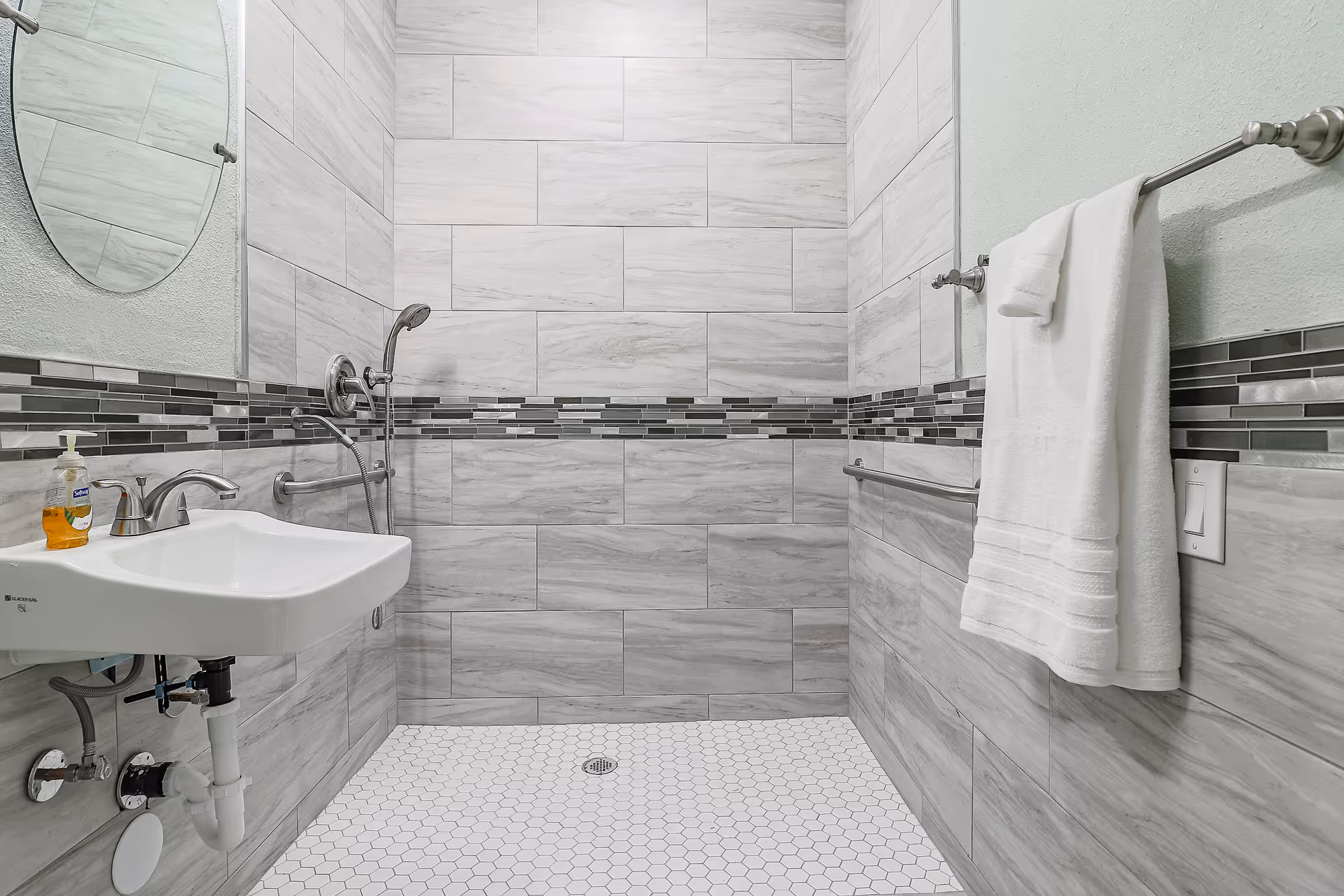 Accessible tiled bathroom with a walk-in shower, wall-mounted sink, grab bars and a towel on a rack.