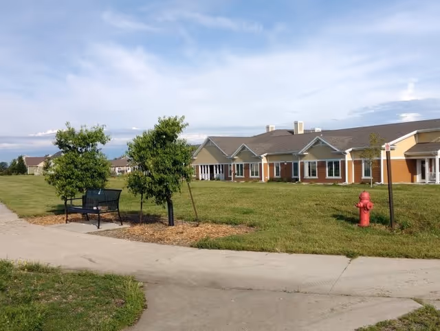 Outdoor view of a senior living facility with a paved walkway, green grass, two small trees, a black bench, a red fire hydrant, and a single-story building with multiple windows and a sloped roof under a partly cloudy sky.