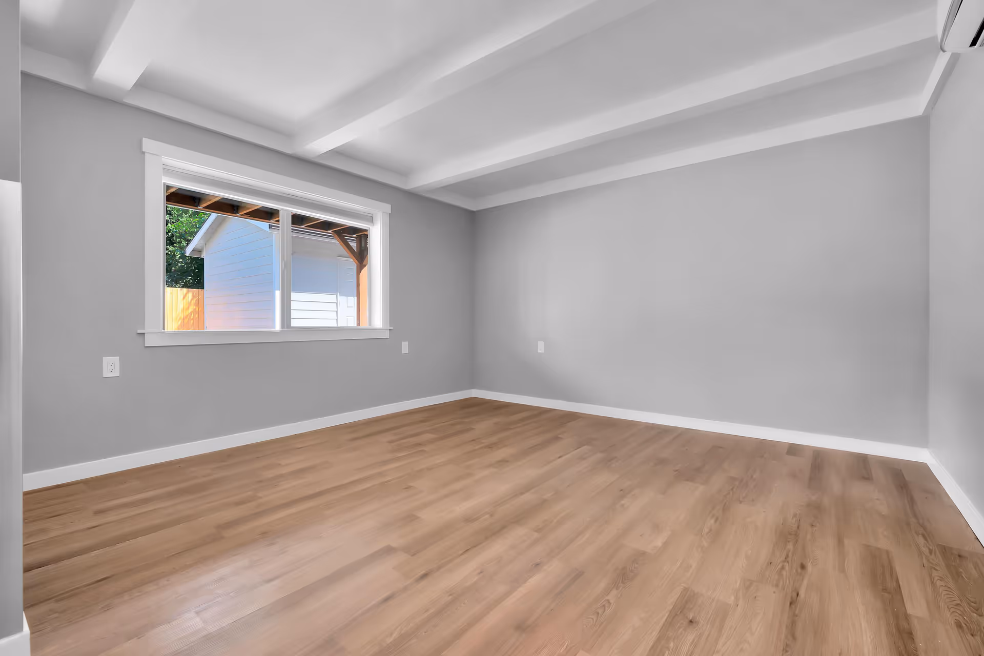 Empty room with light gray walls, wooden floor, a large window showing an outdoor shed, and a white ceiling with exposed beams. An air conditioning unit is mounted on the upper right wall.