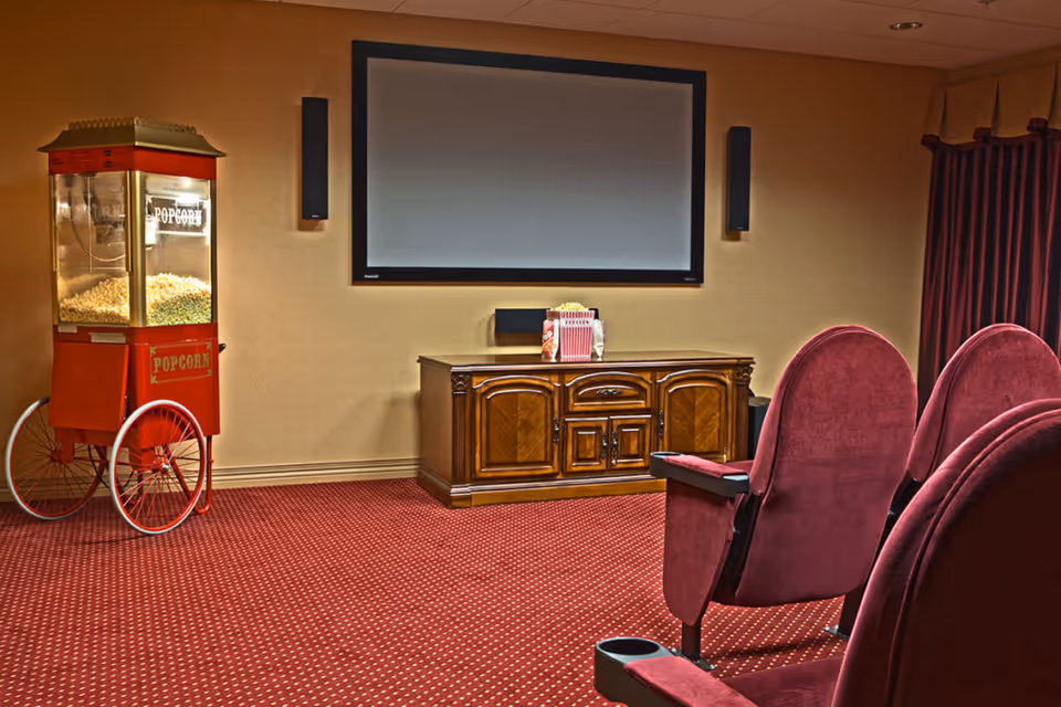 A small theater room with red carpet and red upholstered theater seats facing a large blank projection screen mounted on the wall. To the left, there is a vintage-style popcorn machine filled with popcorn. Below the screen is a wooden cabinet with a popcorn container and some snacks on top.