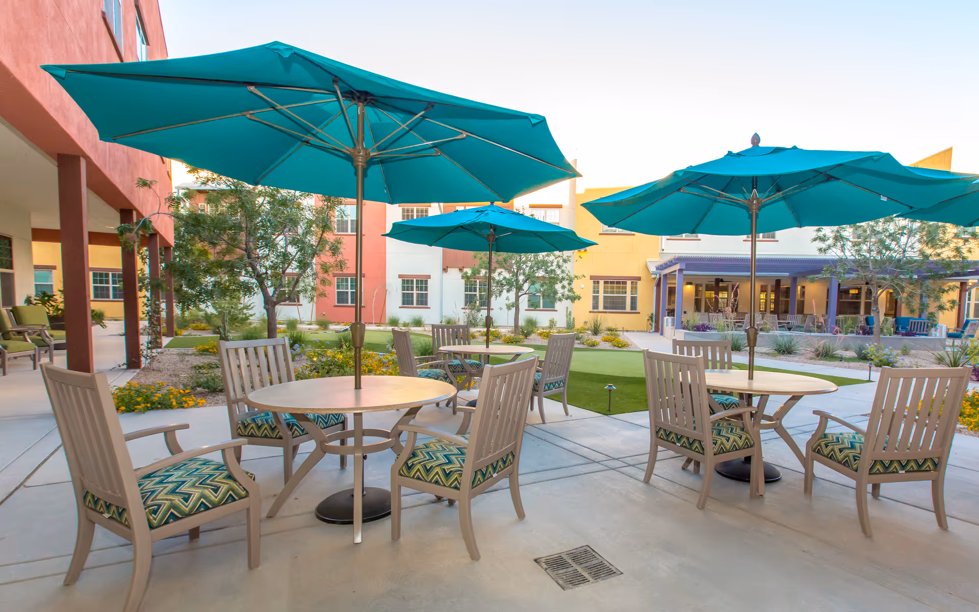 Outdoor courtyard with round tables, teal umbrellas, and chairs in front of a colorful senior living building.