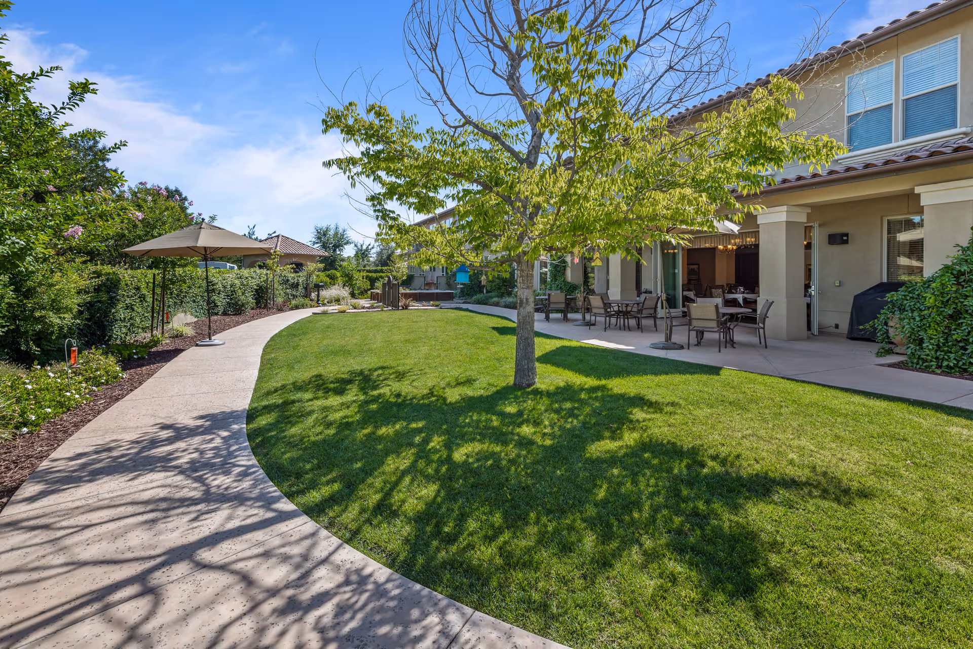 Outdoor area of Oakmont of Fresno featuring a curved concrete pathway, green lawn, a tree casting shadows, patio tables with chairs under a covered porch, and an umbrella providing shade along the path. The building exterior is visible with beige walls and windows.