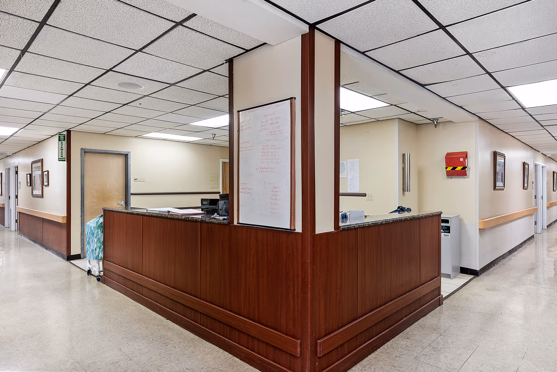 Interior view of a nursing center hallway with a wooden reception desk at the corner. The hallway has beige walls with handrails, framed pictures, and a ceiling with fluorescent lights. There is a whiteboard on the corner wall behind the desk and a fire alarm box on the wall.