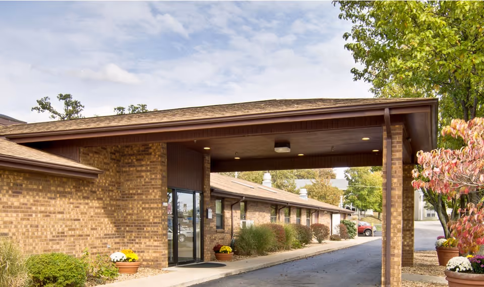Front entrance of a low brick building with a covered drive-through canopy, glass doors, potted plants, and surrounding landscaping.