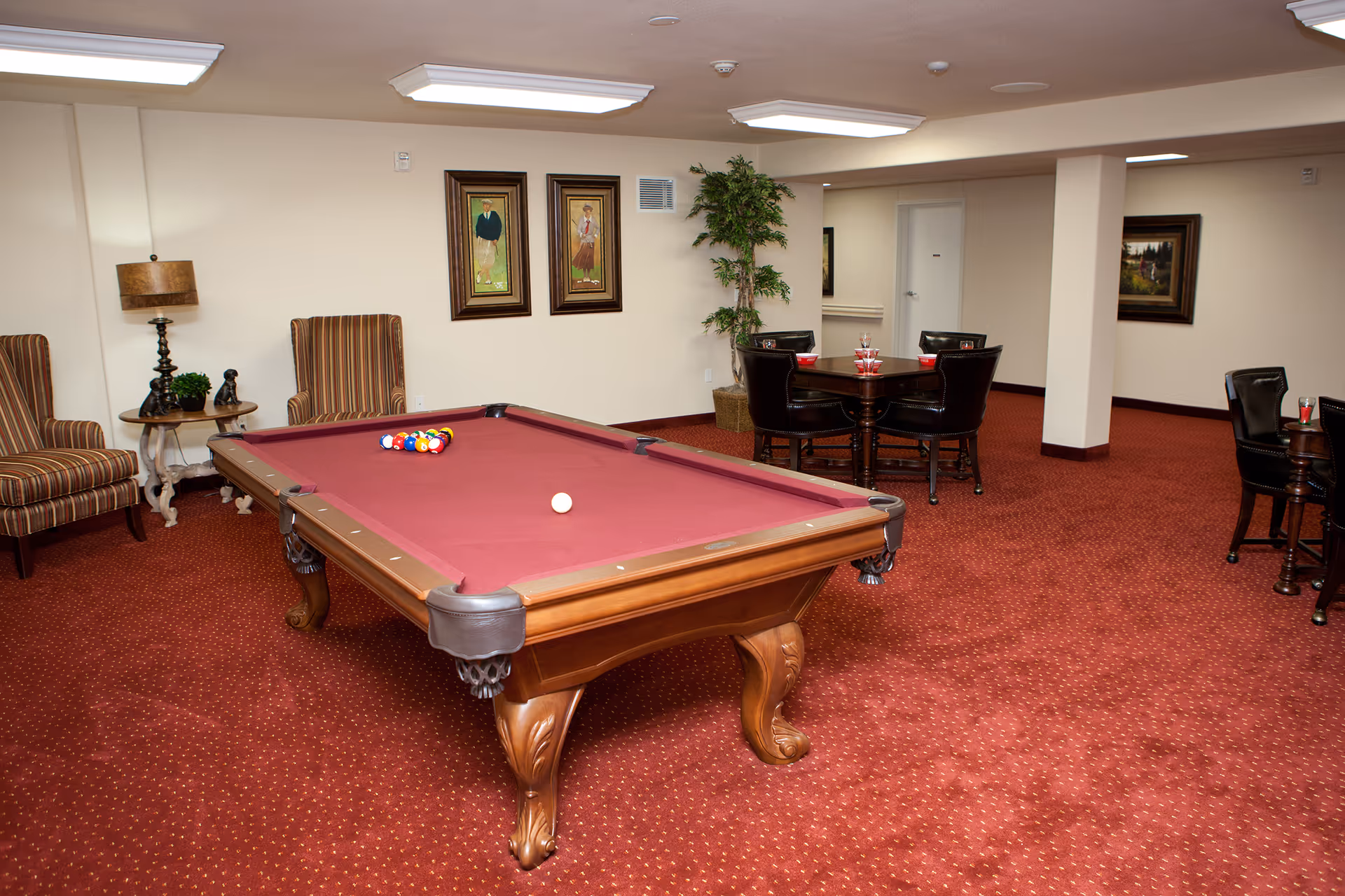 A recreation room with a red felt pool table in the center, surrounded by striped armchairs and a small round table with a lamp and decorative items. In the background, there are two framed paintings on the wall, a potted plant, and a dining table with four black leather chairs set with glasses and bowls. The room has red carpet flooring and beige walls.