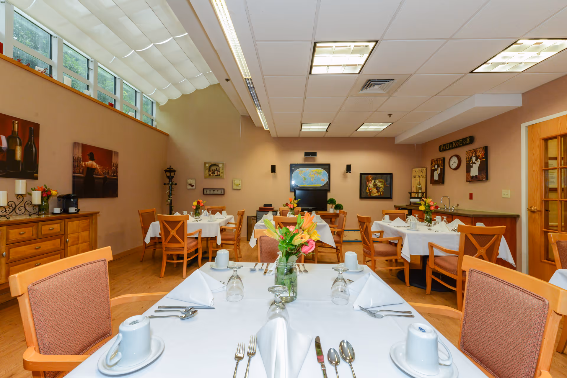 A dining room in an assisted living facility with several tables covered in white tablecloths, each set with folded napkins, cups, glasses, and silverware. There are wooden chairs around the tables and floral centerpieces. The room has beige walls adorned with framed artwork and a sideboard with candles and a coffee maker. Large windows near the ceiling let in natural light.