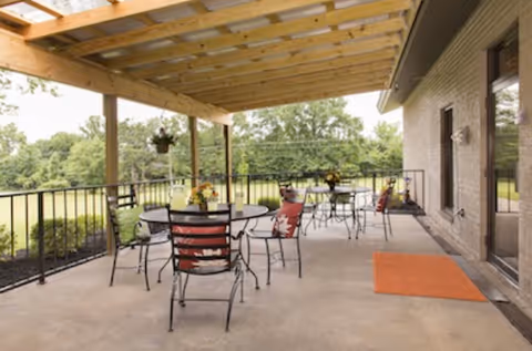 Covered outdoor patio area with several round metal tables and chairs, some with colorful cushions. The patio has a wooden pergola roof and overlooks a green, tree-filled landscape. There is an orange doormat near a door on the right side.