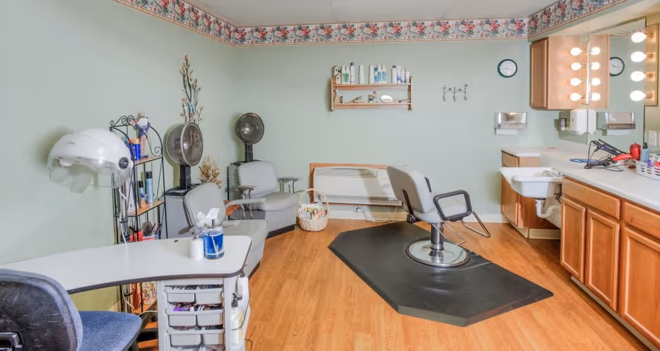Interior view of a salon room in a senior living facility with a salon chair on a black mat, two gray chairs, a hair dryer, a manicure table with supplies, a wall shelf with hair products, a sink with a countertop and cabinets, and a large mirror with lights.