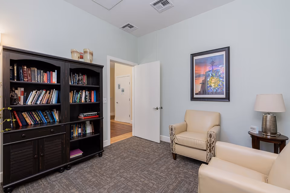 A cozy interior room with two beige armchairs, a dark wooden bookshelf filled with books, a small round wooden side table with a lamp, and a framed colorful artwork on the light blue wall. The door is open, showing a hallway with wooden flooring.
