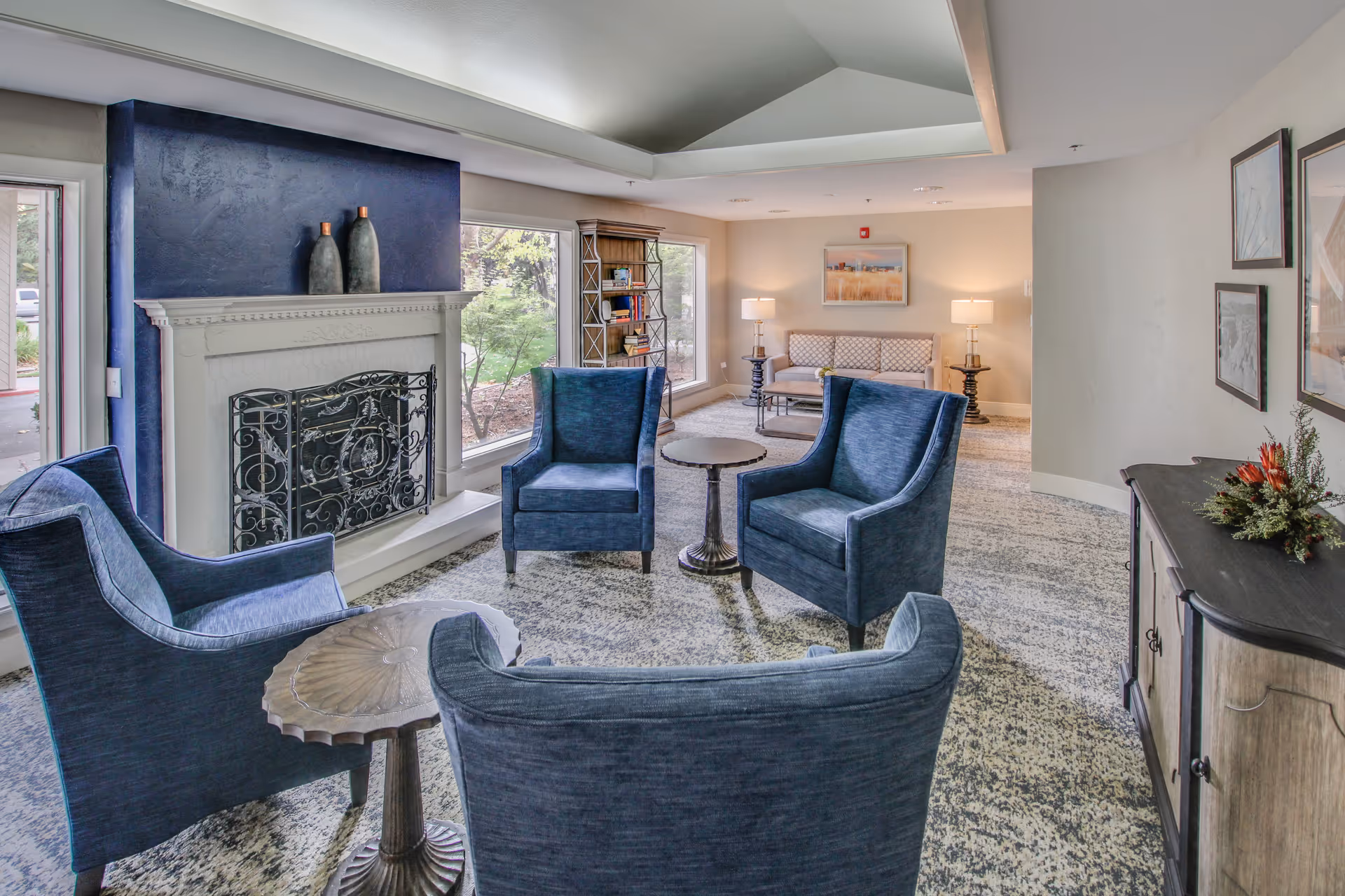 A cozy living room area with four blue upholstered armchairs arranged around two small round tables. There is a white fireplace with a decorative black metal screen and two vases on the mantel. Large windows let in natural light and show greenery outside. In the background, there is a beige sofa with patterned cushions, two table lamps on side tables, a bookshelf, and framed artwork on the walls. The room has a light-colored carpet and neutral walls.