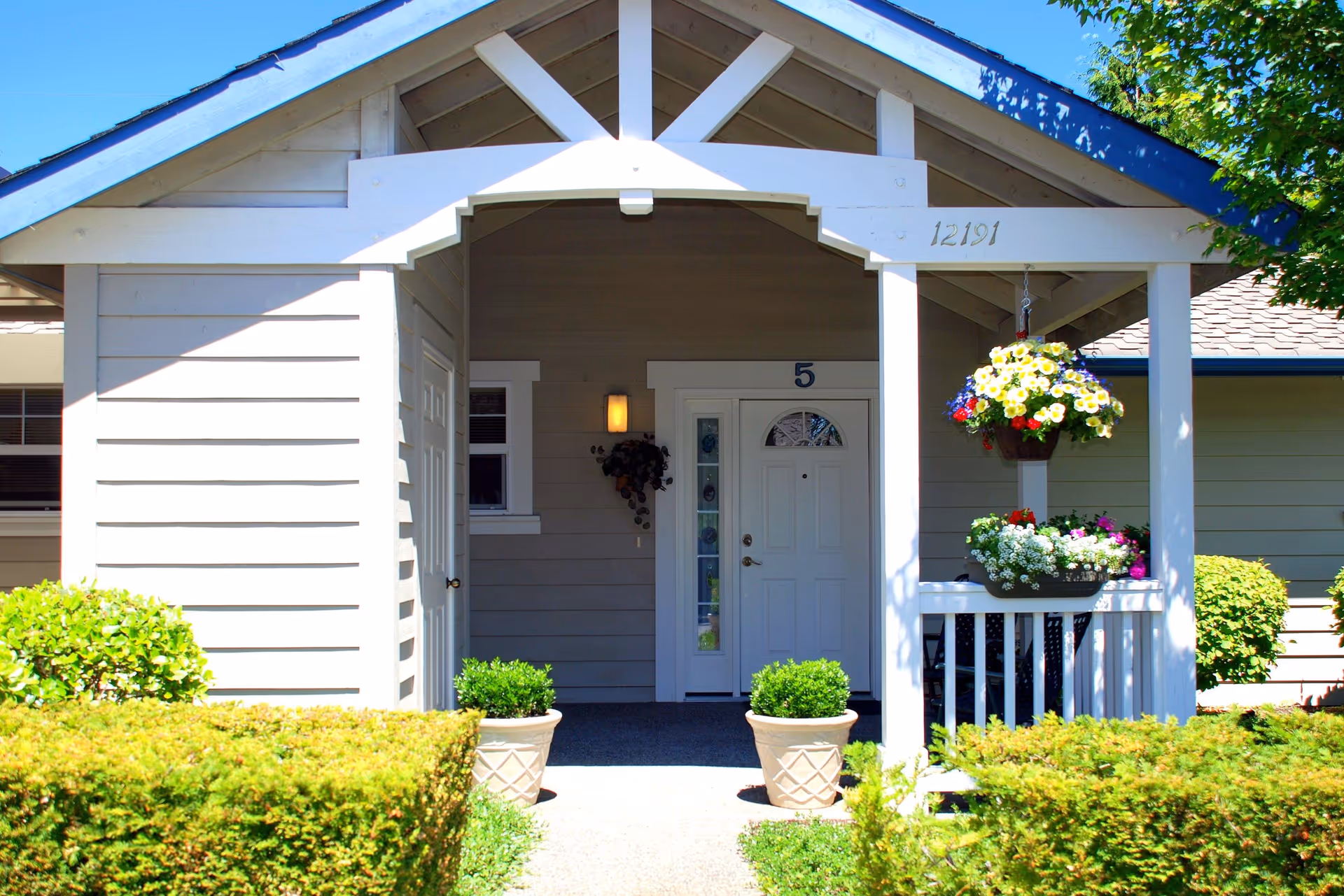 Front porch entrance of a single-story residential building with a white door numbered 5, two potted plants on either side of the walkway, hanging flower baskets, and well-maintained bushes and greenery surrounding the entrance.