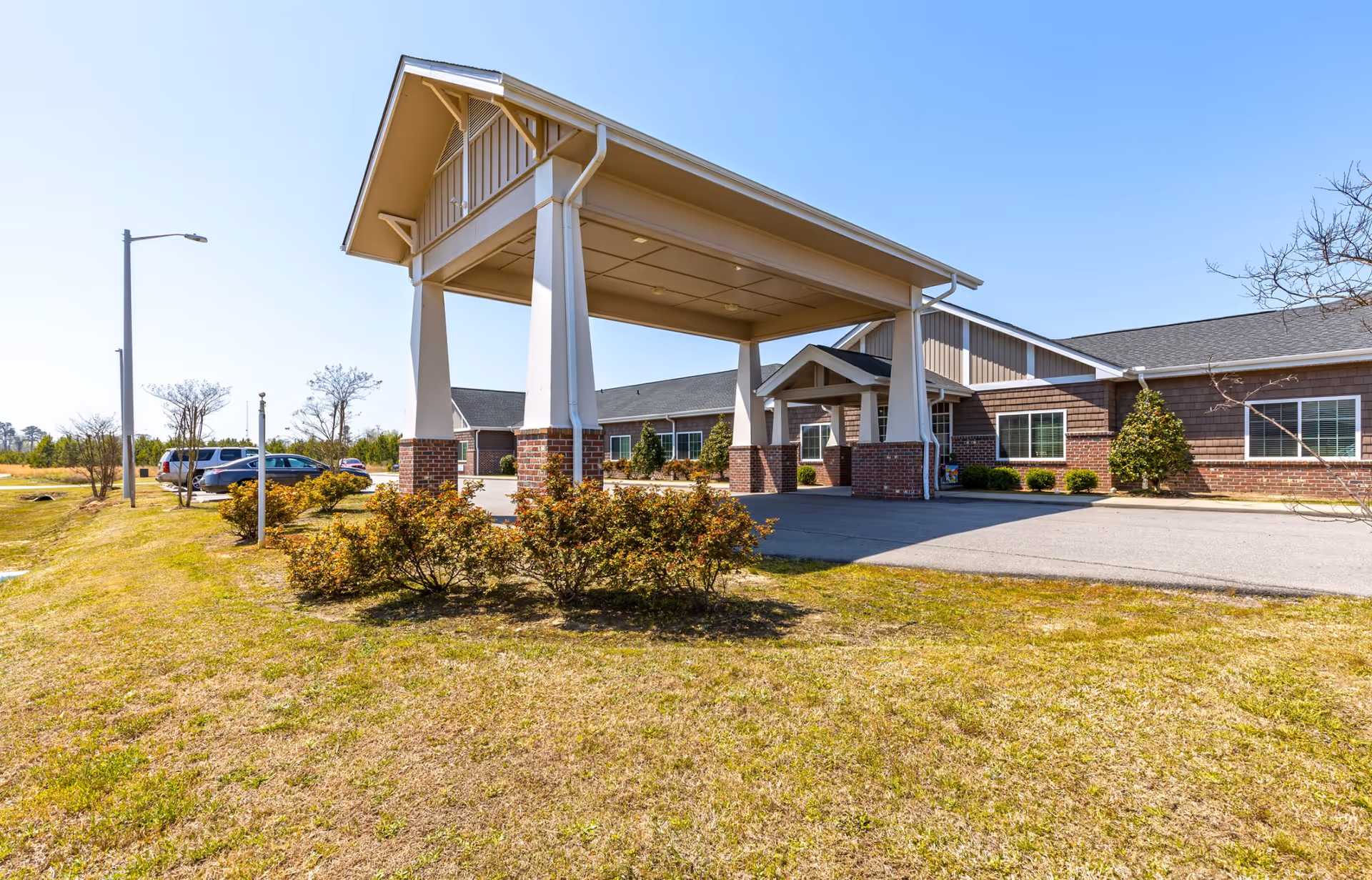 Exterior view of a single-story building with a covered entrance supported by large columns. The building has brick and siding walls with several windows. There are bushes and a grassy area in the foreground, a few parked cars to the left, and a clear blue sky above.