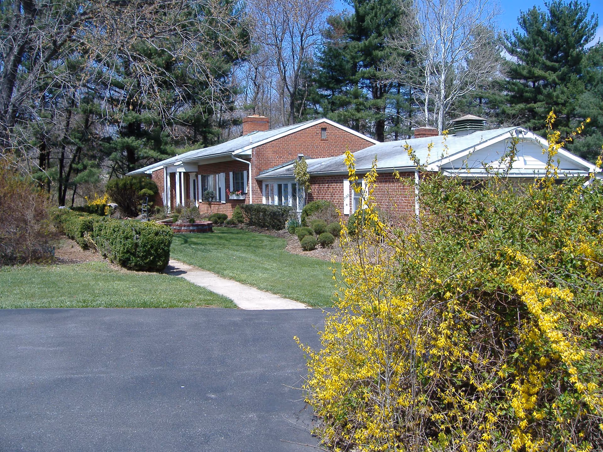 A single-story brick building with white trim surrounded by green grass, bushes, and trees. There is a paved driveway in the foreground and a concrete walkway leading to the building entrance. Yellow flowering bushes are visible on the right side of the image under a clear blue sky.