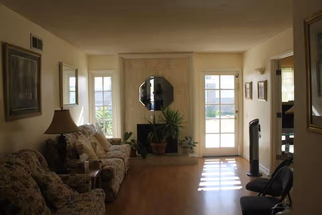A cozy living room with floral patterned sofas, a wooden side table with a lamp, a fireplace with a decorative mirror above it, several potted plants, and a glass door letting in natural light. The room has light-colored walls and wooden flooring.