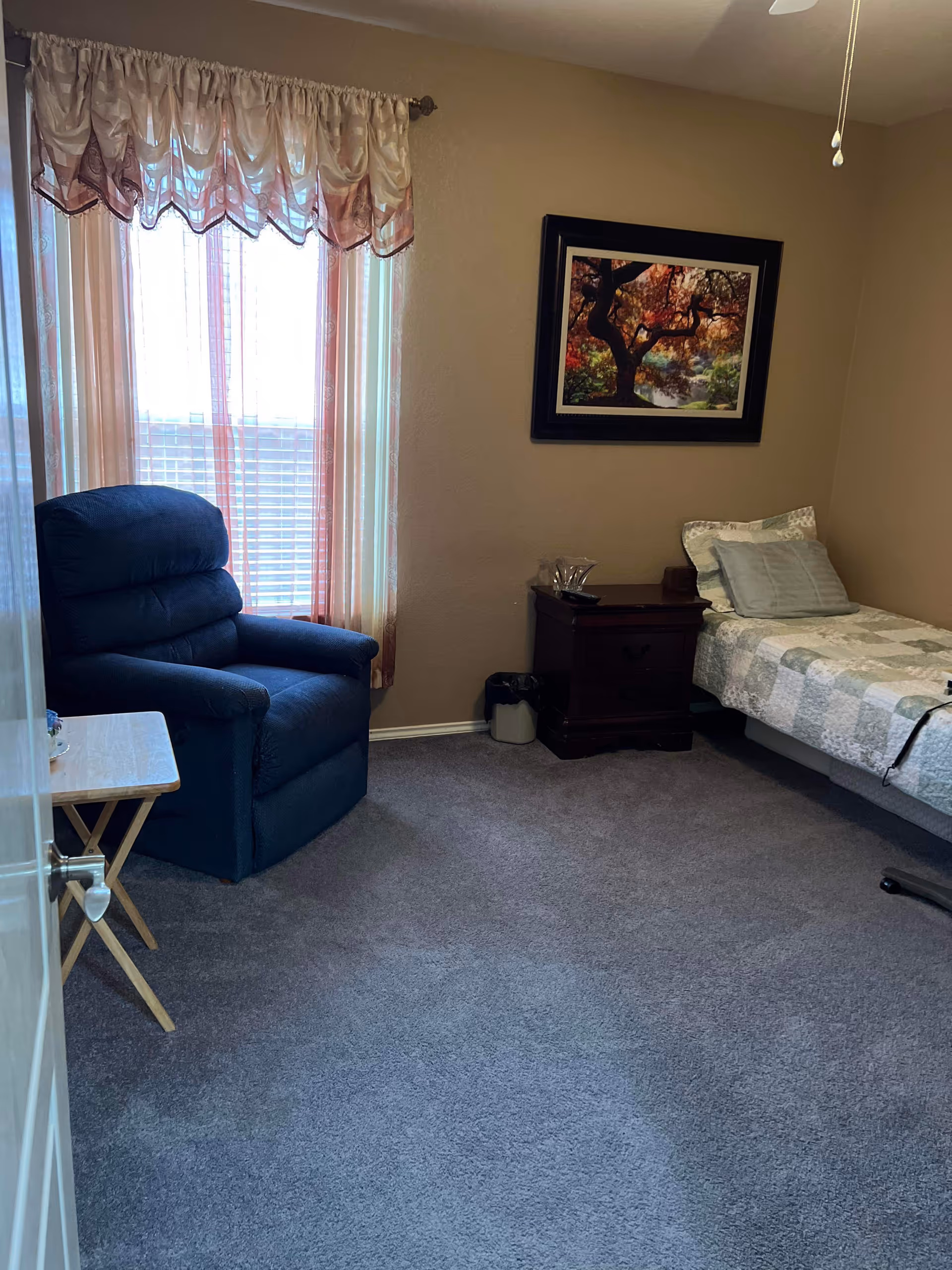A small bedroom with a single bed covered in a patchwork quilt, a wooden nightstand with a glass pitcher and cup, a blue recliner chair, a small folding table, and a window with sheer and decorative curtains. A framed picture of a tree with autumn leaves hangs on the wall above the nightstand.