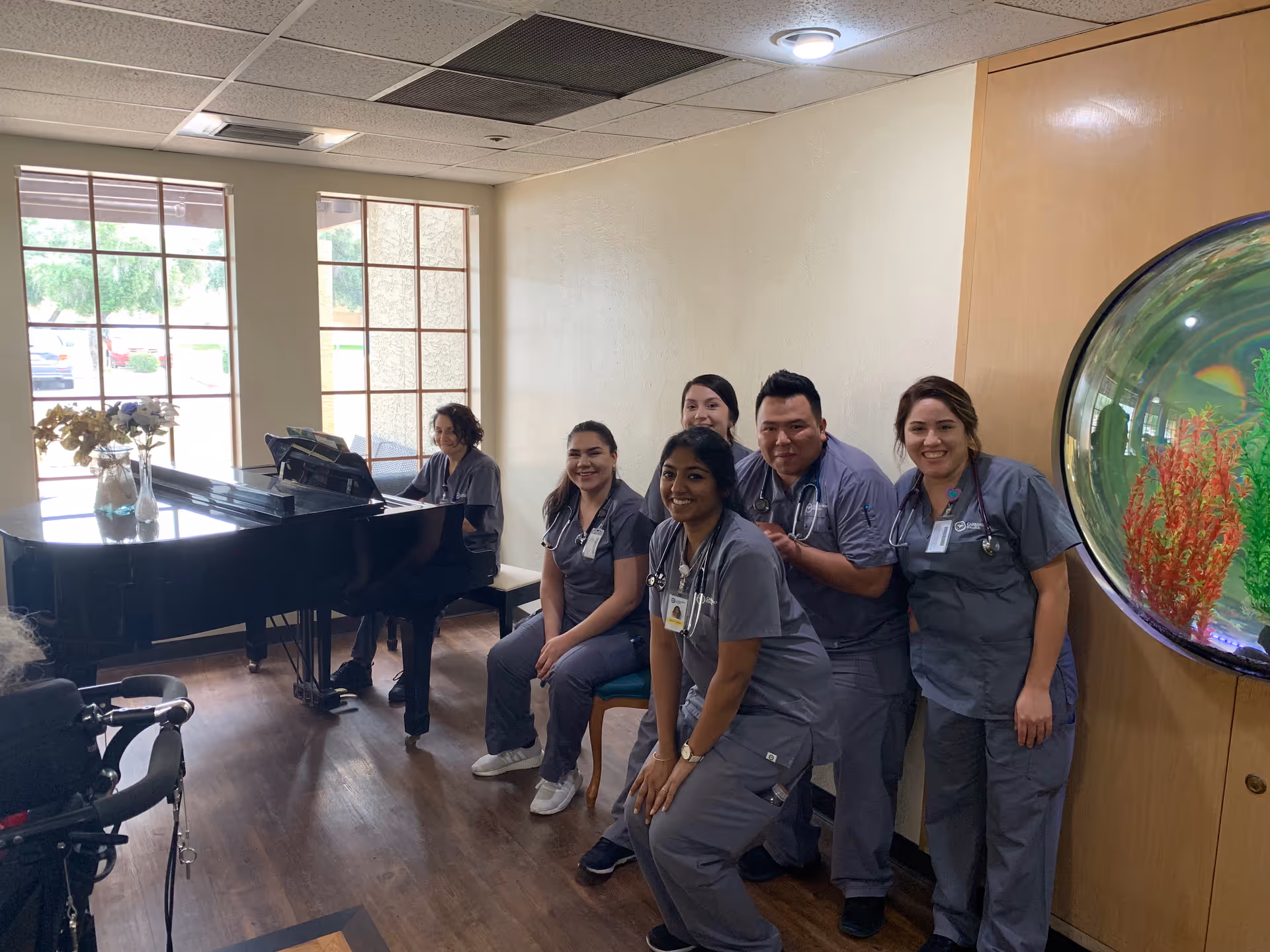 A group of five healthcare workers in gray scrubs posing and smiling inside a room with wooden floors and large windows. One person is seated at a black grand piano, and there is a large round fish tank mounted on the wall to the right. A wheelchair is partially visible in the foreground.