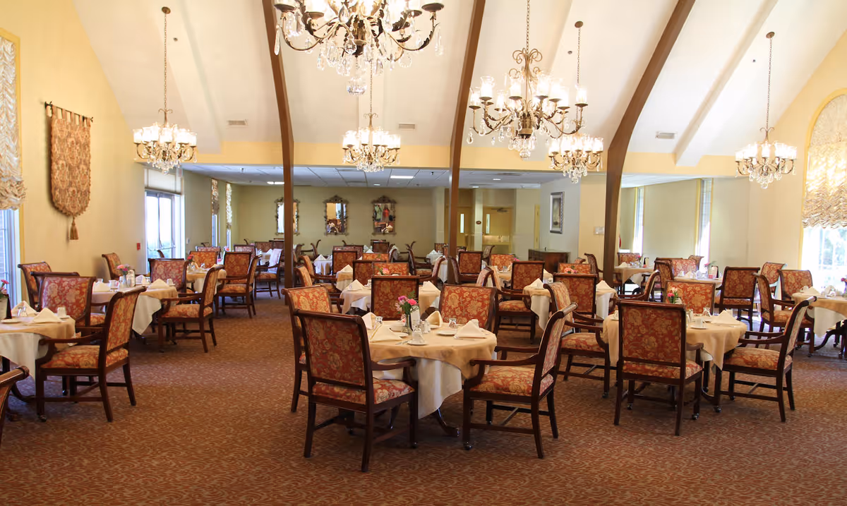 A spacious dining room with multiple round tables covered with white tablecloths, each set with napkins, plates, and glassware. The chairs have a floral upholstery pattern. The room features several ornate chandeliers hanging from a high ceiling with wooden beams. Large windows with decorative curtains allow natural light to fill the space.