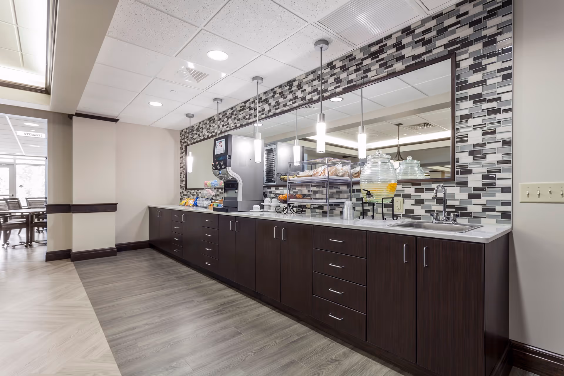 A modern kitchenette area with dark wood cabinets, a white countertop, a sink, a beverage dispenser with lemonade, a coffee machine, and a display case with pastries. The backsplash features a mosaic tile design in shades of gray, white, and black. Three pendant lights hang above the counter, and a large mirror runs along the wall behind the counter. In the background, there is a dining area with tables and chairs.