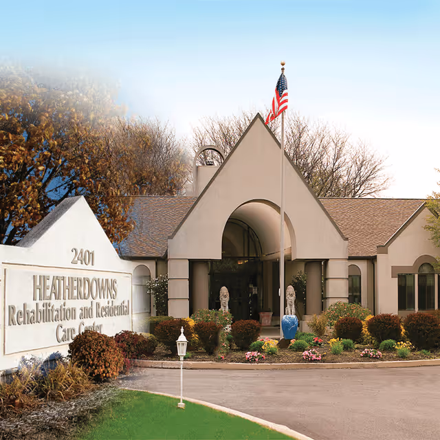 Exterior view of Heatherdowns Rehabilitation and Residential Care Center building with a peaked roof, an American flag on a flagpole in front, landscaped bushes and flowers, and a large sign displaying the facility name and address 2401.