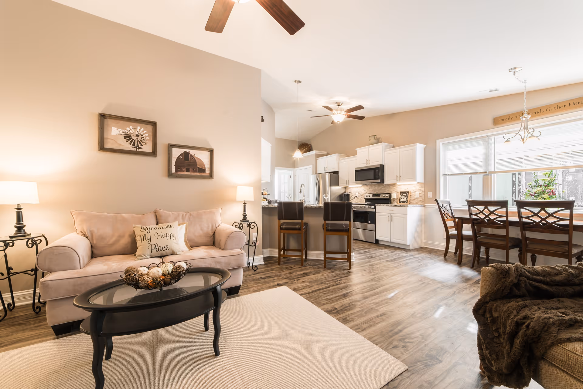 A bright and cozy open-concept living space featuring a beige couch with a decorative pillow that reads 'Sycamore My Happy Place', two side tables with lamps, a black oval coffee table with a decorative centerpiece, a kitchen area with white cabinets, stainless steel appliances, and two bar stools at a granite countertop, and a dining area with a wooden table and chairs near large windows letting in natural light.