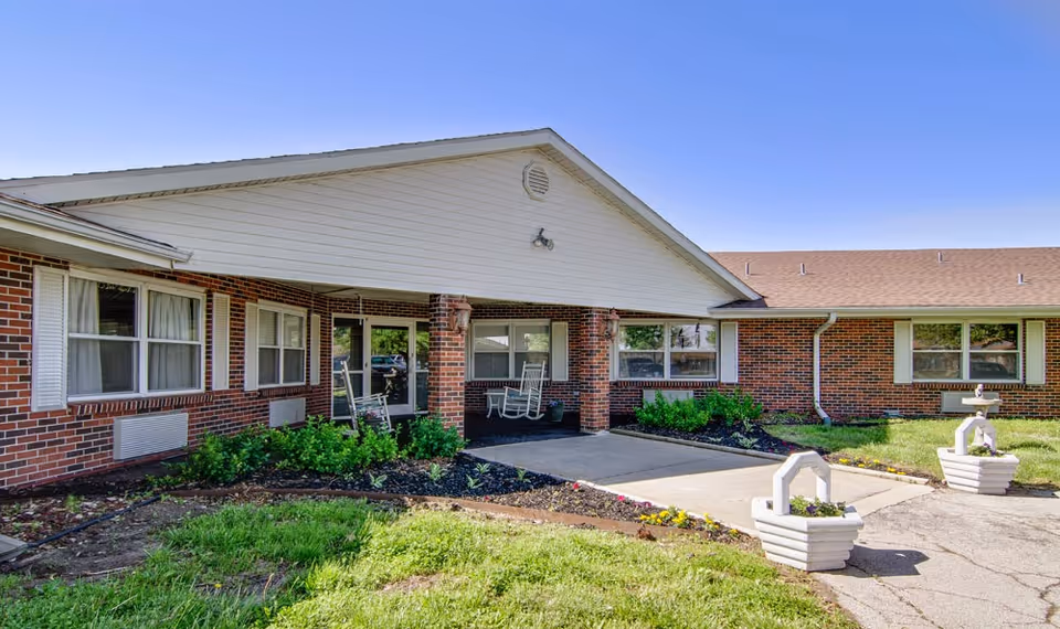 Exterior view of a single-story brick building with a covered entrance supported by brick columns. There are two white rocking chairs on the porch, green shrubs along the building, and two white planters with flowers near the driveway. The sky is clear and blue.