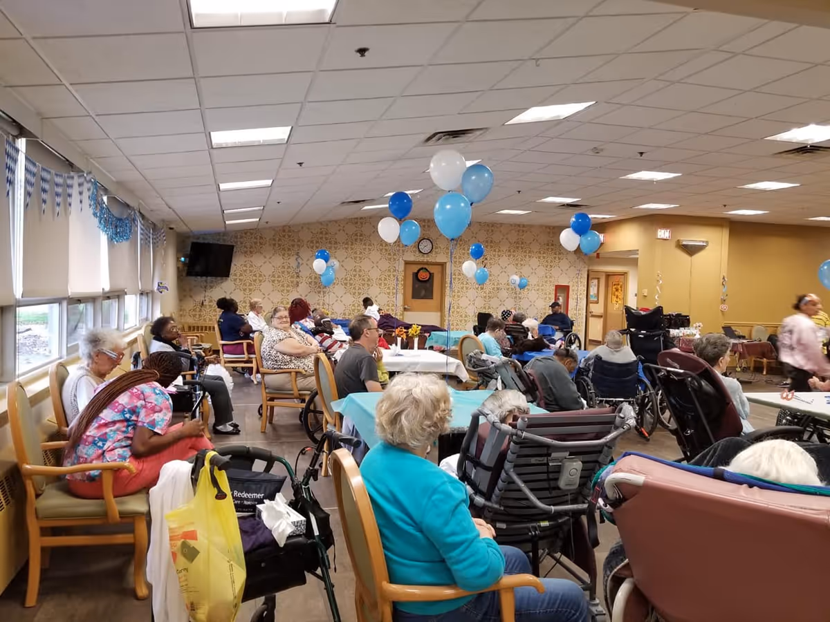 Large decorated common/dining room in a senior living facility with residents seated at tables and balloons overhead.