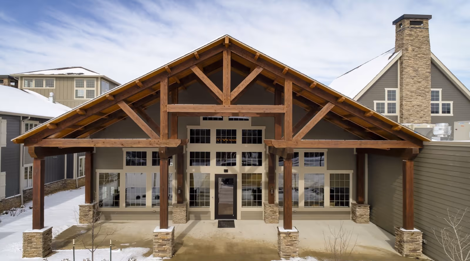 Front entrance of a senior living building with a large exposed-wood gabled porch, stone pillars, and snow on the ground and rooftops.