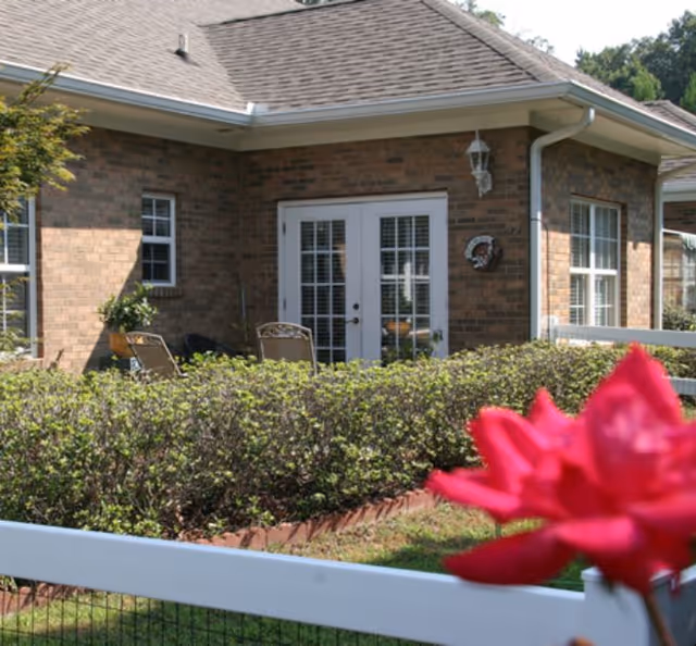 View of a brick building with white-framed French doors and windows, surrounded by green bushes and a white fence. A red flower is prominently visible in the foreground on the right side.