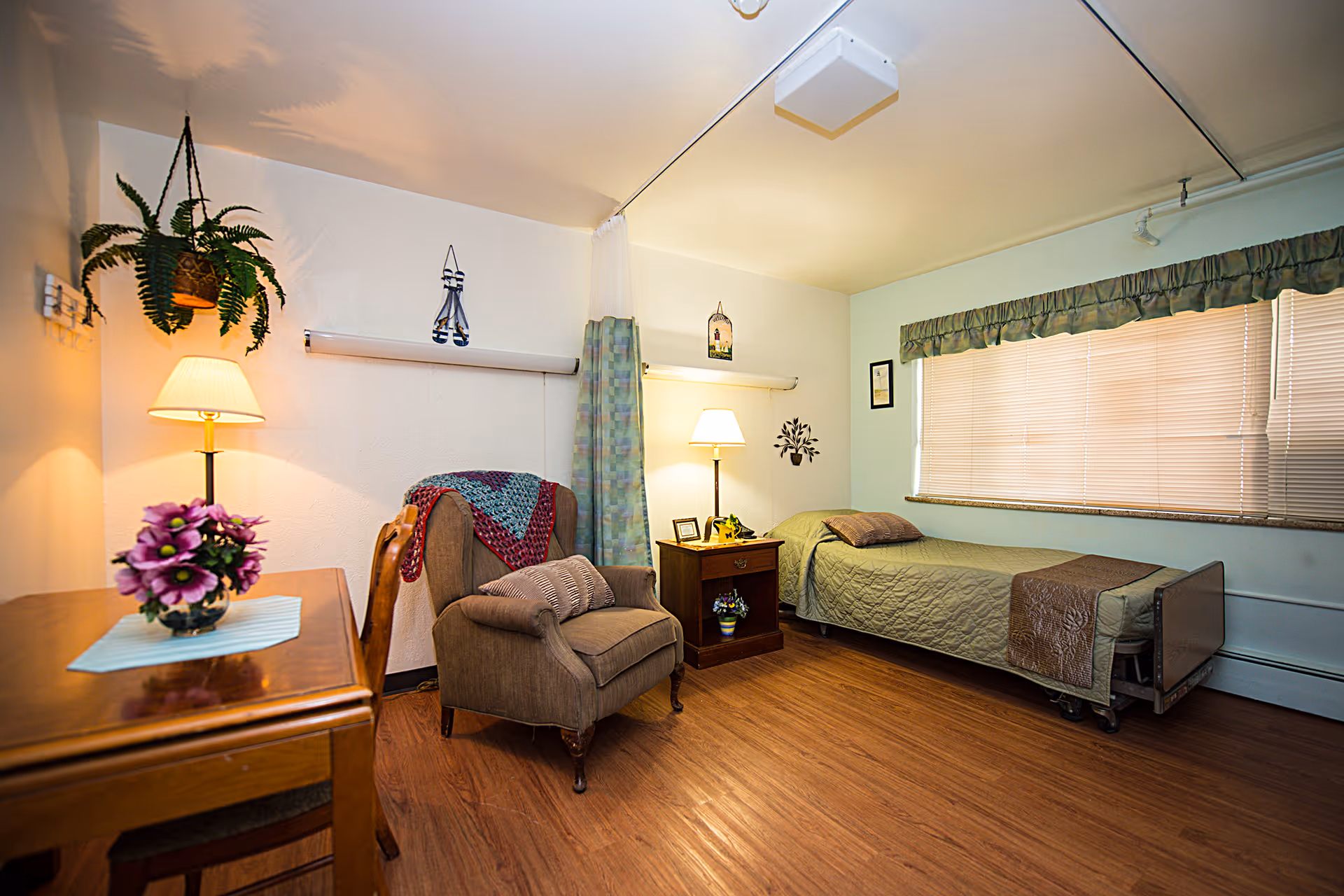 A cozy nursing center room with a single bed covered in a green quilt and a brown throw, a wooden nightstand with a lamp and small decorations, a comfortable armchair with a cushion and a crocheted blanket draped over it, a wooden table with a vase of purple flowers, a hanging plant, and a large window with closed blinds and a valance.