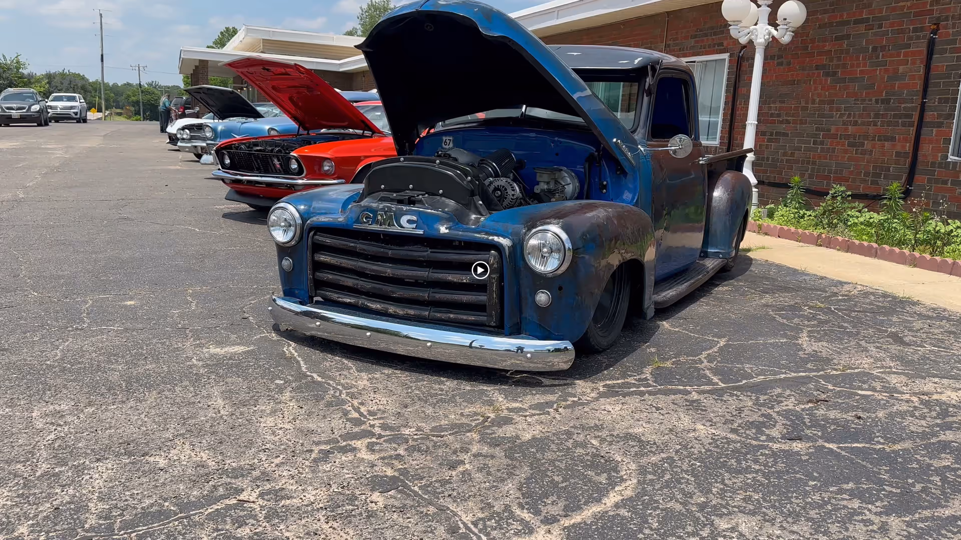 A row of vintage cars with their hoods open parked on an asphalt surface outside a brick building with a white lamp post and some greenery.