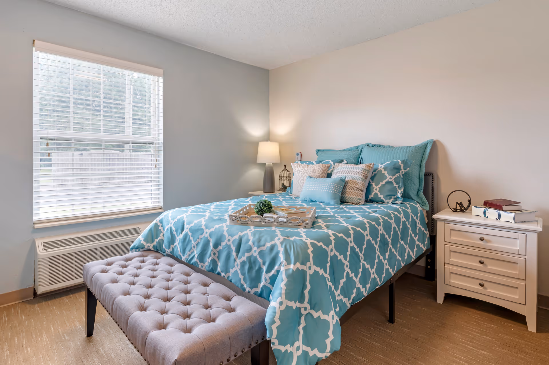 A neatly made bed with a blue and white patterned comforter and multiple pillows in a bedroom. There is a cushioned bench at the foot of the bed, a window with blinds on the left wall, and a white nightstand with books and decorative items on the right side. A lamp is turned on beside the bed.