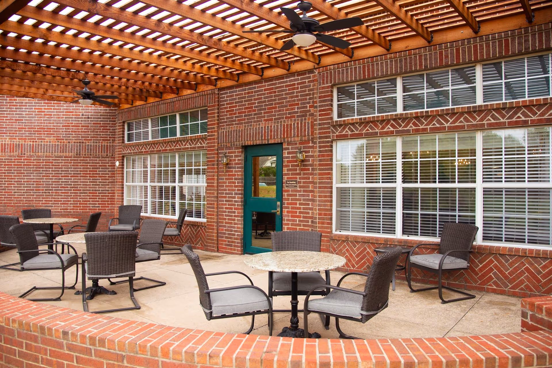 Outdoor patio area with a wooden pergola overhead, several round tables with cushioned chairs arranged on a concrete floor, and a brick wall with large windows and a green door in the background.