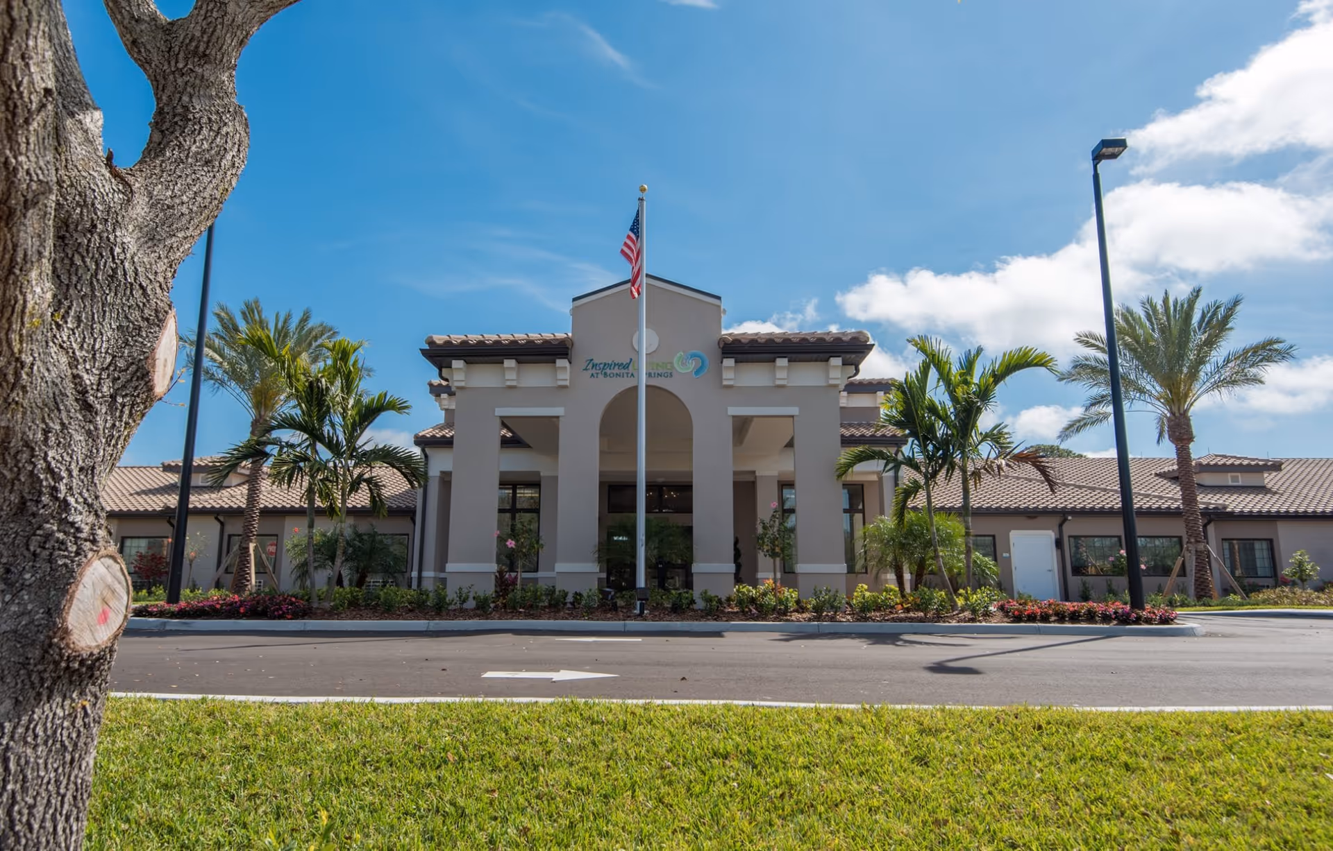 Front entrance of a Mediterranean-style senior living building with an American flag, palm trees, and landscaped grounds.