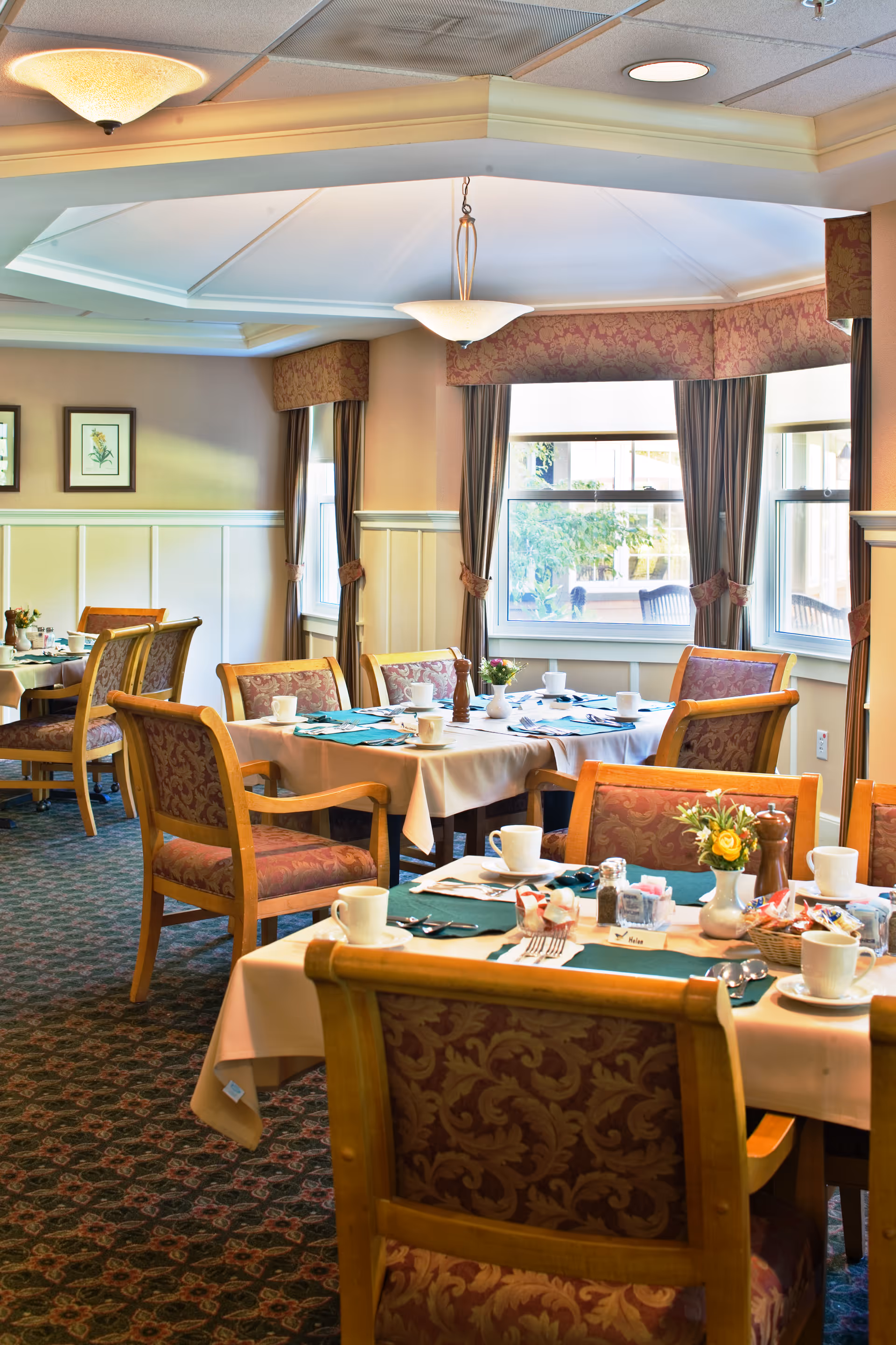 A well-lit dining room with several tables covered in white tablecloths and green placemats. Each table is set with white cups, plates, silverware, and small flower arrangements. The room has large windows with curtains, patterned carpet, and upholstered wooden chairs with a floral design.