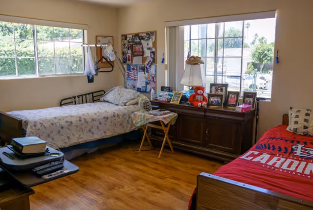 A furnished senior living bedroom with two beds, dresser, bulletin board, and personal photos.