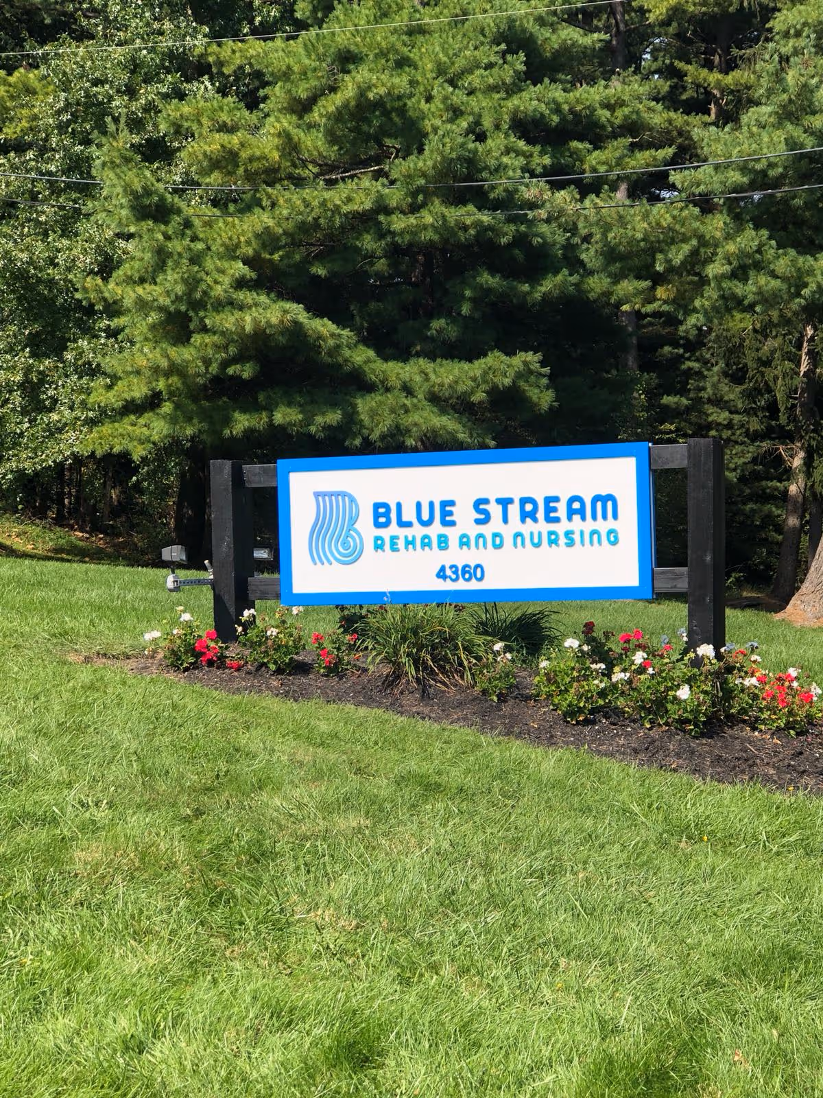 A sign for Blue Stream Rehab and Nursing with the address number 4360, set in a landscaped area with green grass, flowers, and trees in the background.