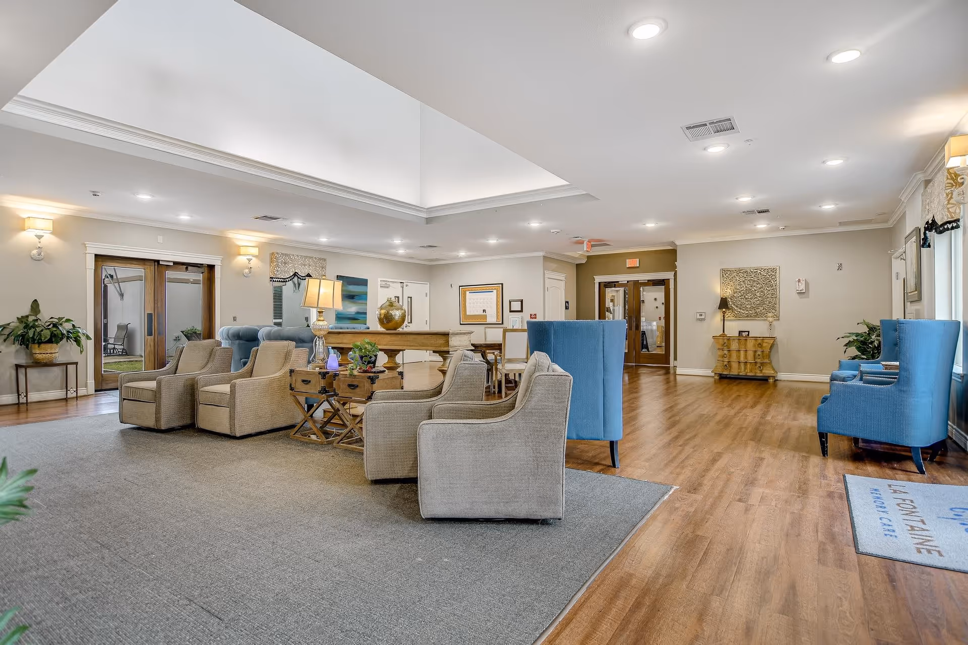 A spacious and well-lit common area in La Fontaine Memory Care featuring a seating arrangement with beige and blue upholstered chairs around wooden tables. The room has hardwood floors, a large gray area rug, decorative plants, wall art, and multiple light fixtures on the ceiling and walls. There are double doors leading to an outdoor area and another set of double doors at the far end of the room.