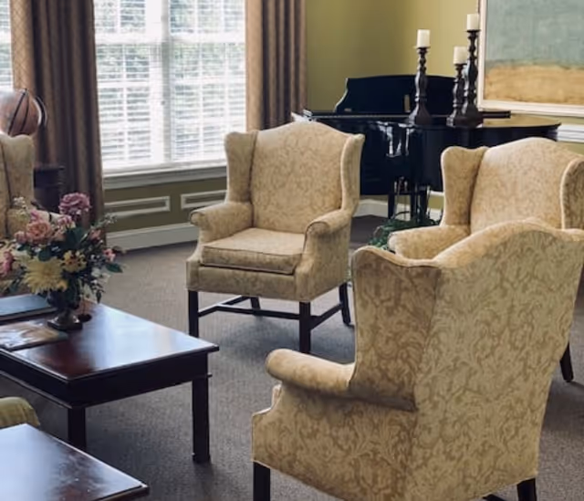 A cozy living room area with four beige patterned armchairs arranged around a dark wooden coffee table. There is a bouquet of flowers on the table. In the background, a large window with white blinds and brown curtains lets in natural light. A black grand piano with several white candles on top is positioned against a green wall.
