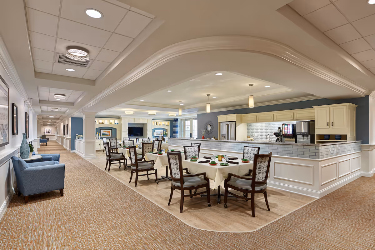 Bright open dining area in an assisted living facility with neatly set tables, wooden chairs, a serving counter and a long adjacent hallway with seating.