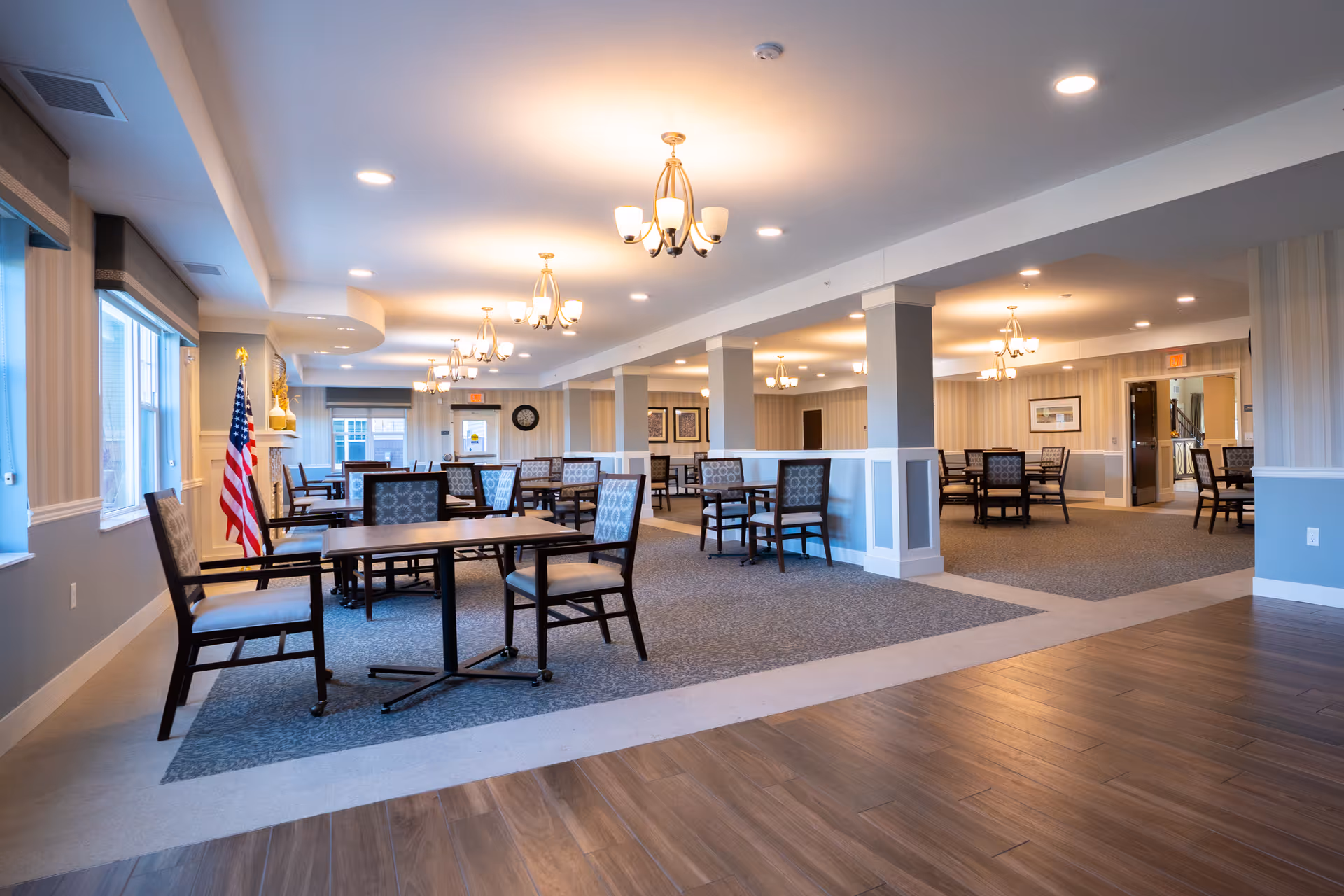 Spacious dining area in a senior living facility with multiple tables and chairs arranged neatly. The room features large windows on the left side, allowing natural light to fill the space. There are several chandeliers hanging from the ceiling, and an American flag is positioned near the windows. The flooring is a combination of carpet and wood, and the walls are decorated with framed artwork and a clock.