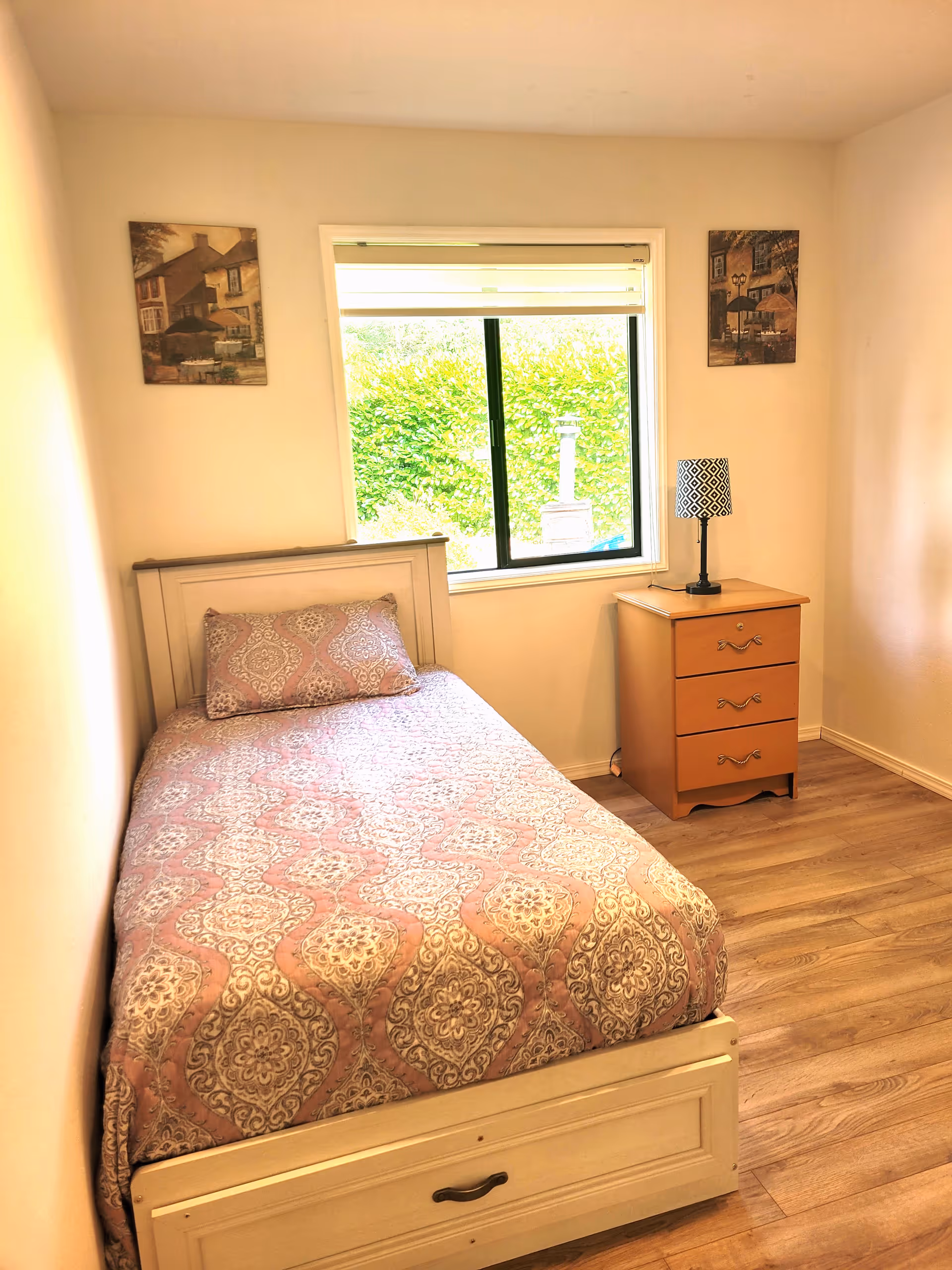A small bedroom with a single bed covered in a patterned pink and beige bedspread and matching pillow. There is a wooden nightstand with three drawers next to the bed, topped with a black and white patterned lamp. A window with a white blind is centered on the wall behind the nightstand, showing greenery outside. Two framed pictures hang on the wall, one on each side of the window. The room has light-colored walls and wood flooring.