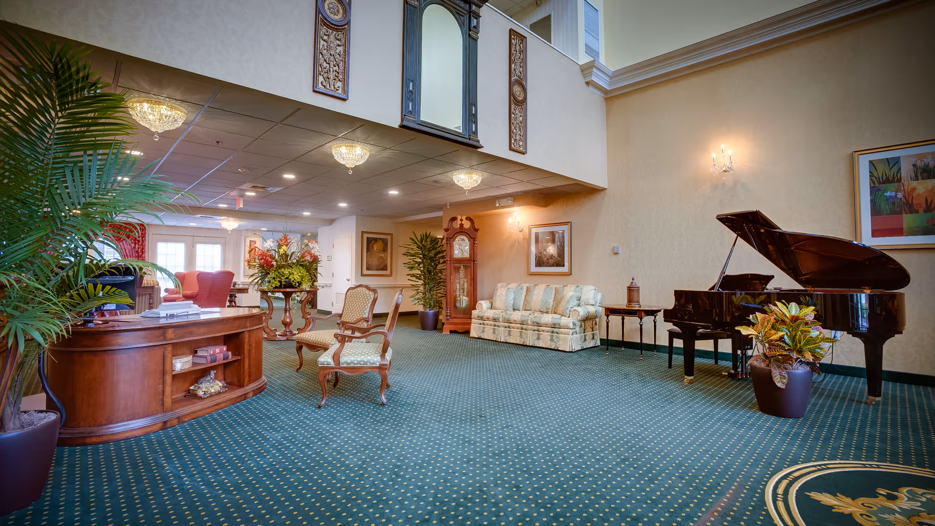 A spacious and elegant living room area with green patterned carpet, a grand piano, a floral sofa, wooden chairs, a wooden reception desk, potted plants, a grandfather clock, framed artwork on the walls, and decorative ceiling lights.
