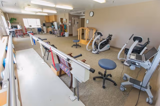 A rehabilitation room with parallel bars for walking exercises, exercise bikes, stools, and various therapy equipment. The room has beige walls, carpeted floor, and fluorescent lighting. There is a clock on the wall and a window letting in natural light.
