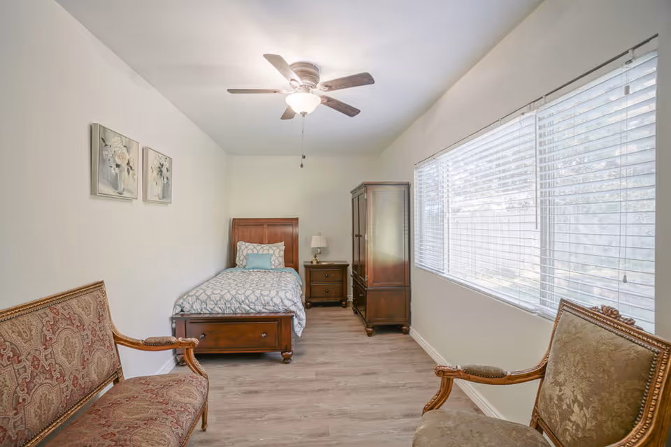 A bright bedroom with a single wooden bed with patterned bedding, a wooden nightstand with a lamp, a wooden wardrobe, two upholstered chairs with ornate wooden frames, and two floral paintings on the wall. A large window with blinds lets in natural light.