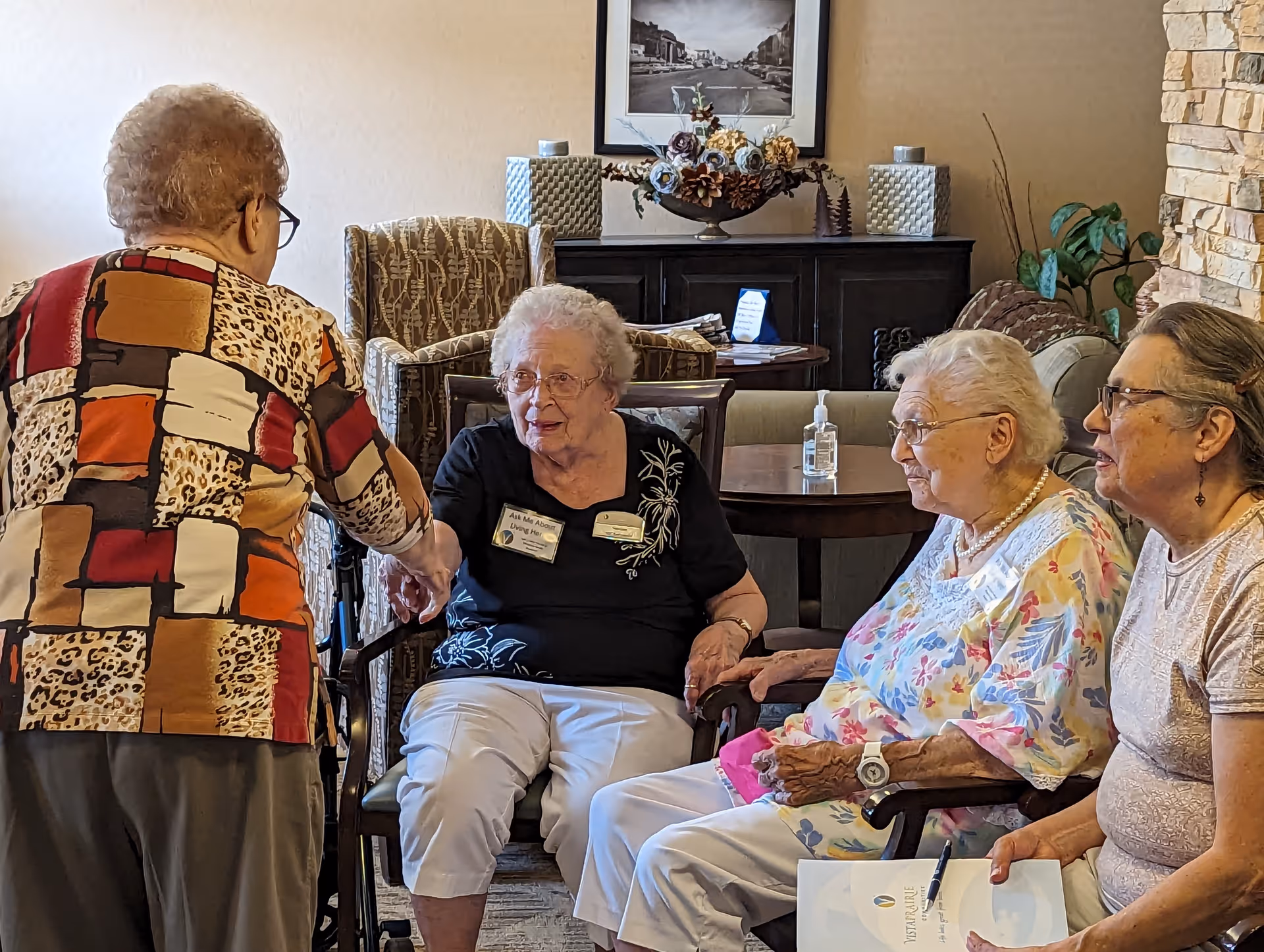 Four elderly women interacting in a cozy living room setting. Three women are seated, two holding hands, while the fourth woman stands and shakes hands with one of the seated women. The room has comfortable chairs, a table with a hand sanitizer bottle, decorative flowers, and a framed picture on the wall.