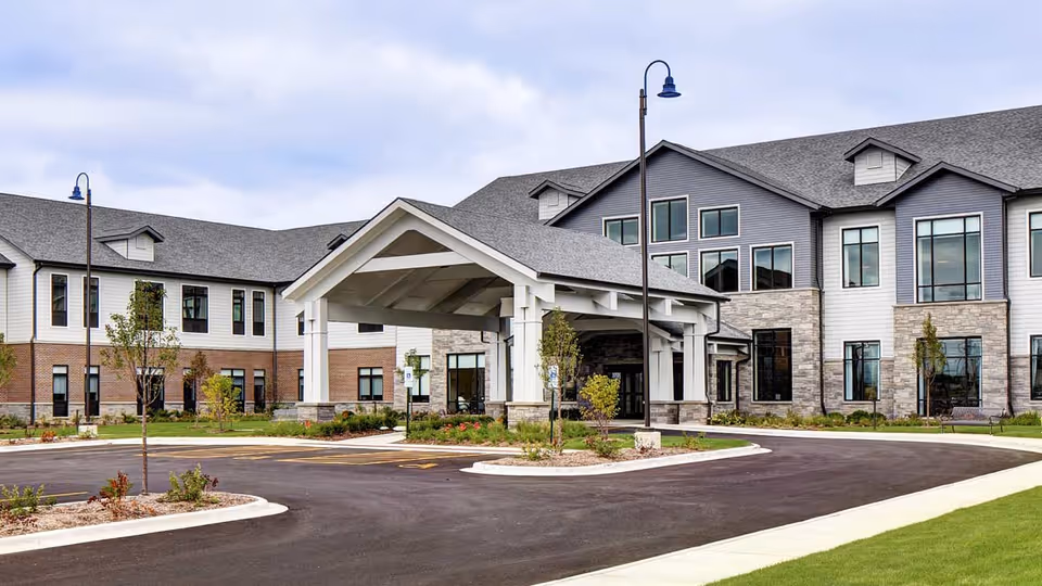 Exterior view of a modern senior living facility building with a covered entrance, multiple windows, and a well-maintained driveway and landscaping under a cloudy sky.