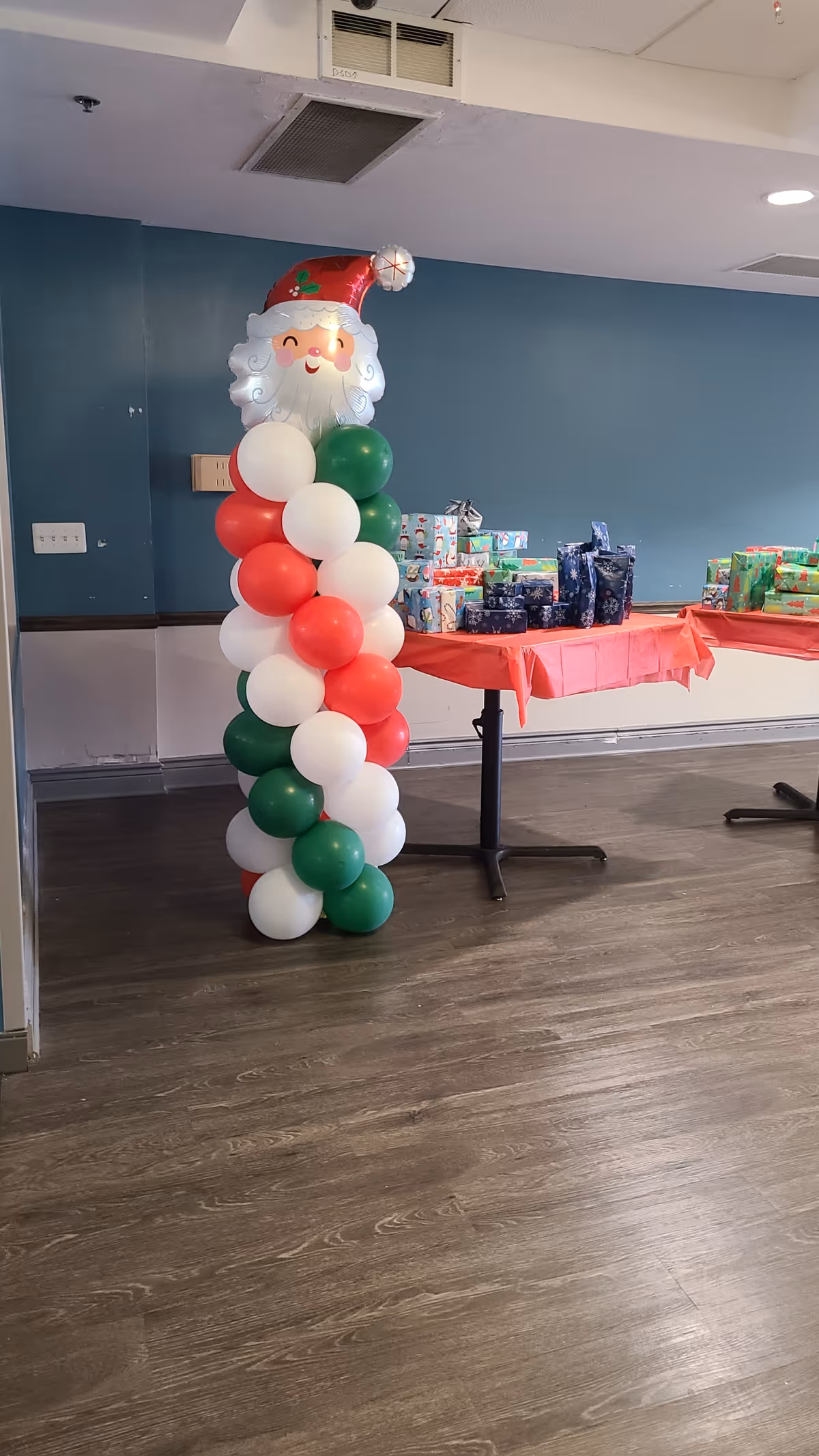 A room with wood flooring and teal walls featuring a tall balloon decoration shaped like Santa Claus made of red, white, and green balloons. Two tables covered with red tablecloths hold wrapped Christmas presents.