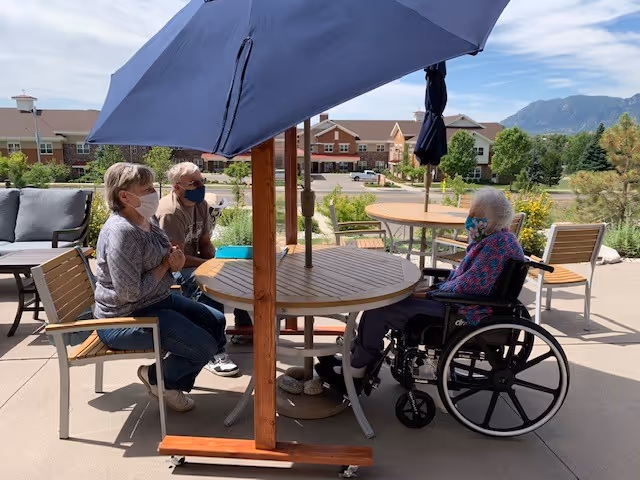 Three elderly people sitting outdoors at a round wooden table with a large blue umbrella. Two people are seated on chairs, and one person is in a wheelchair. All are wearing face masks. The background shows a senior living facility building, greenery, and mountains under a partly cloudy sky.