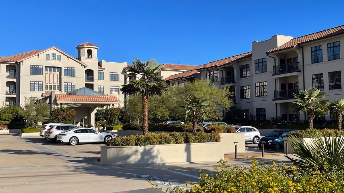 Front exterior of a multi-story Mediterranean-style senior living building with parked cars, palm trees, and a driveway under a clear blue sky.